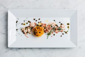 Vit rektangulär tallrik med tunnskivad bläckfisk, grillad citron, kapris och färska örter på marmorbord.