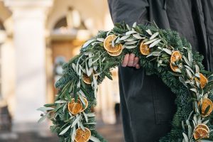 Hand håller en julkrans dekorerad med torkade apelsinskivor och gröna kvistar framför Villa Aske.