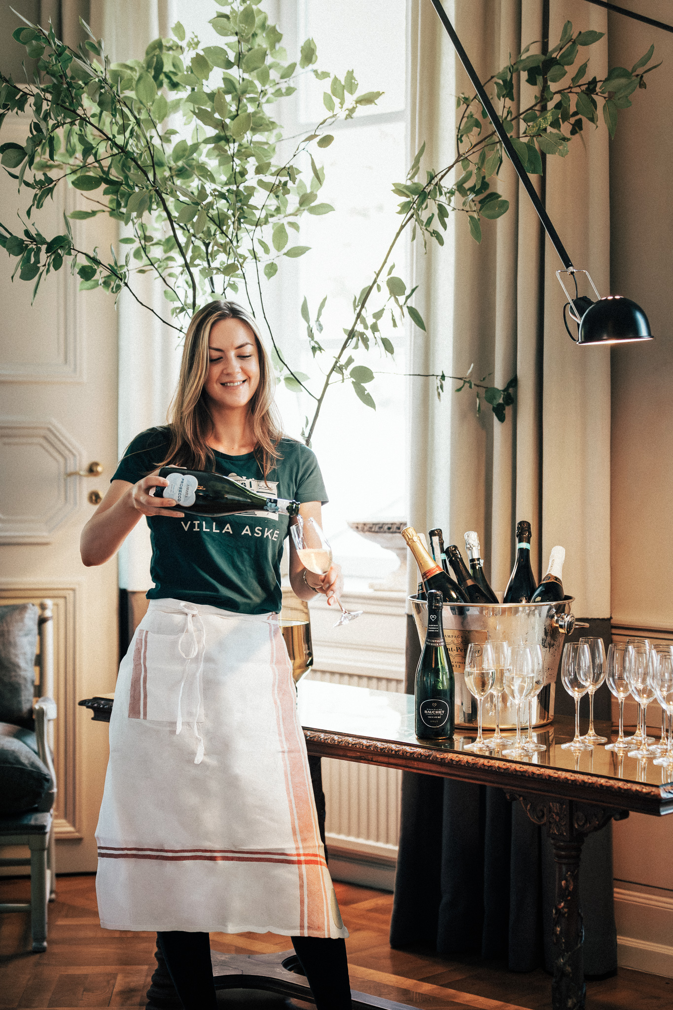 Servitris på Villa Aske häller upp mousserande vin i glas vid ett bord dukat med champagneflaskor och glas.